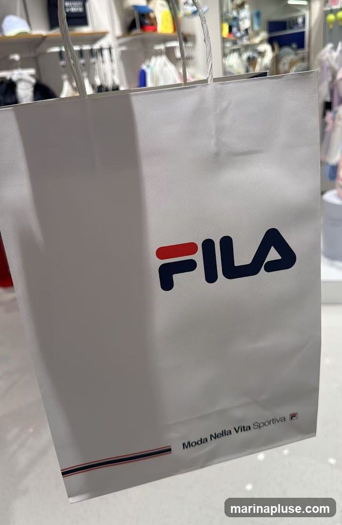 A white shopping bag with the Fila logo and the text "Moda Nella Vita Sportiva" printed on it. The background shows a retail store interior with clothing on display racks.