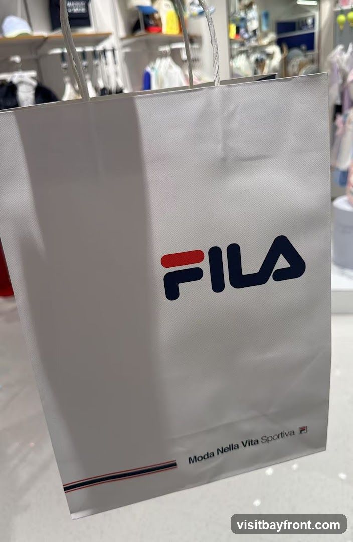 A white shopping bag with the Fila logo and the text "Moda Nella Vita Sportiva" printed on it. The background shows a retail store interior with clothing on display racks.