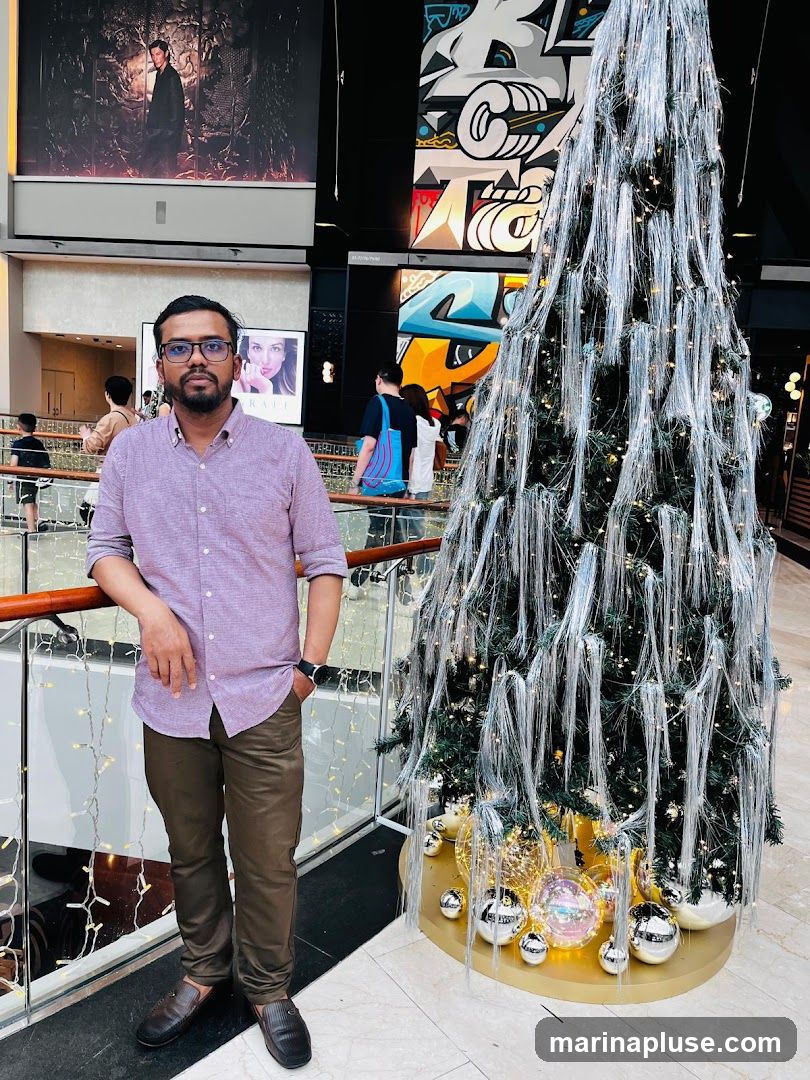 A man stands in a well-lit indoor space, possibly a mall, next to a tall, elaborately decorated Christmas tree. The tree is adorned with silver tinsel-like strands and small lights, with large, reflective ornaments at its base. Behind the man, a large mural with graffiti-style art is visible on the wall. Further in the background, other shoppers can be seen, and large advertisements are displayed on the walls. The man is wearing a light purple collared shirt and brown pants.
