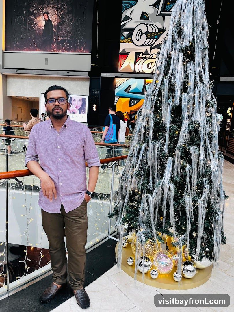 A man stands in a well-lit indoor space, possibly a mall, next to a tall, elaborately decorated Christmas tree. The tree is adorned with silver tinsel-like strands and small lights, with large, reflective ornaments at its base. Behind the man, a large mural with graffiti-style art is visible on the wall. Further in the background, other shoppers can be seen, and large advertisements are displayed on the walls. The man is wearing a light purple collared shirt and brown pants.