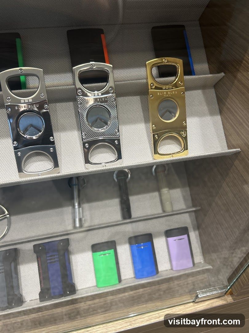 A display case featuring various cigar cutters and lighters. The top shelf shows three Elie Bleu cigar cutters in silver, silver with black accents, and gold. Behind them are dark cases with colored accents (green, orange, blue). The middle shelf displays several lighters. The bottom shelf showcases colorful lighters in green, blue, and purple, along with other dark-colored lighters.