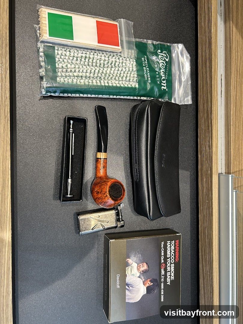 A collection of pipe smoking accessories laid out on a dark surface. Items include a briar pipe, a metal lighter, a pipe tool, a black leather pipe case, a package of Peterson pipe cleaners with brush, and a box of Davidoff cigarettes with a health warning label. A small packet with what appears to be Italian flag stickers is also visible.
