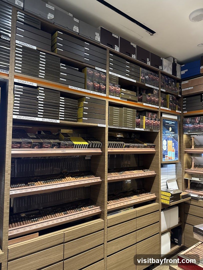 A well-organized display of cigars in a store. Shelves are stocked with numerous boxes and individual cigars, categorized by brand and type. The wooden shelving units and drawers suggest a humidor environment. A poster is visible on the right side, advertising cigars.