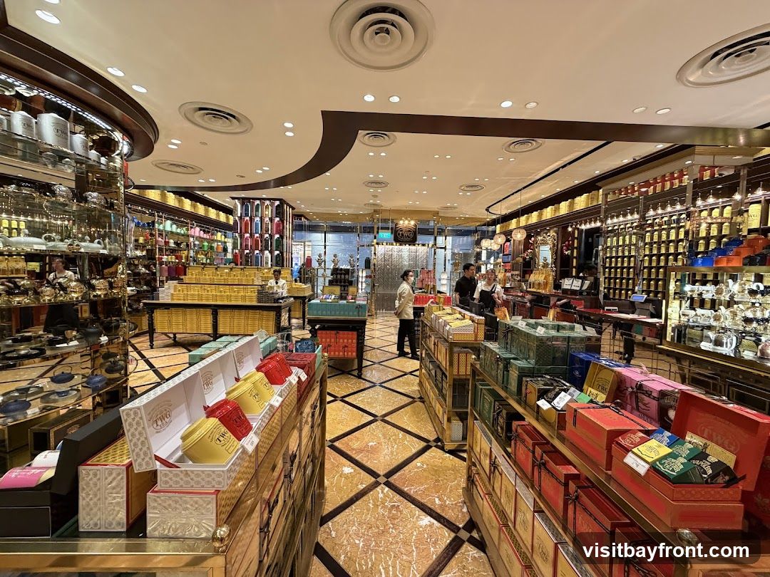 The interior of a well-lit tea shop, showcasing rows of shelves stocked with various tea tins and gift boxes. The floor is a marble pattern with gold accents. Several staff members are visible assisting customers.