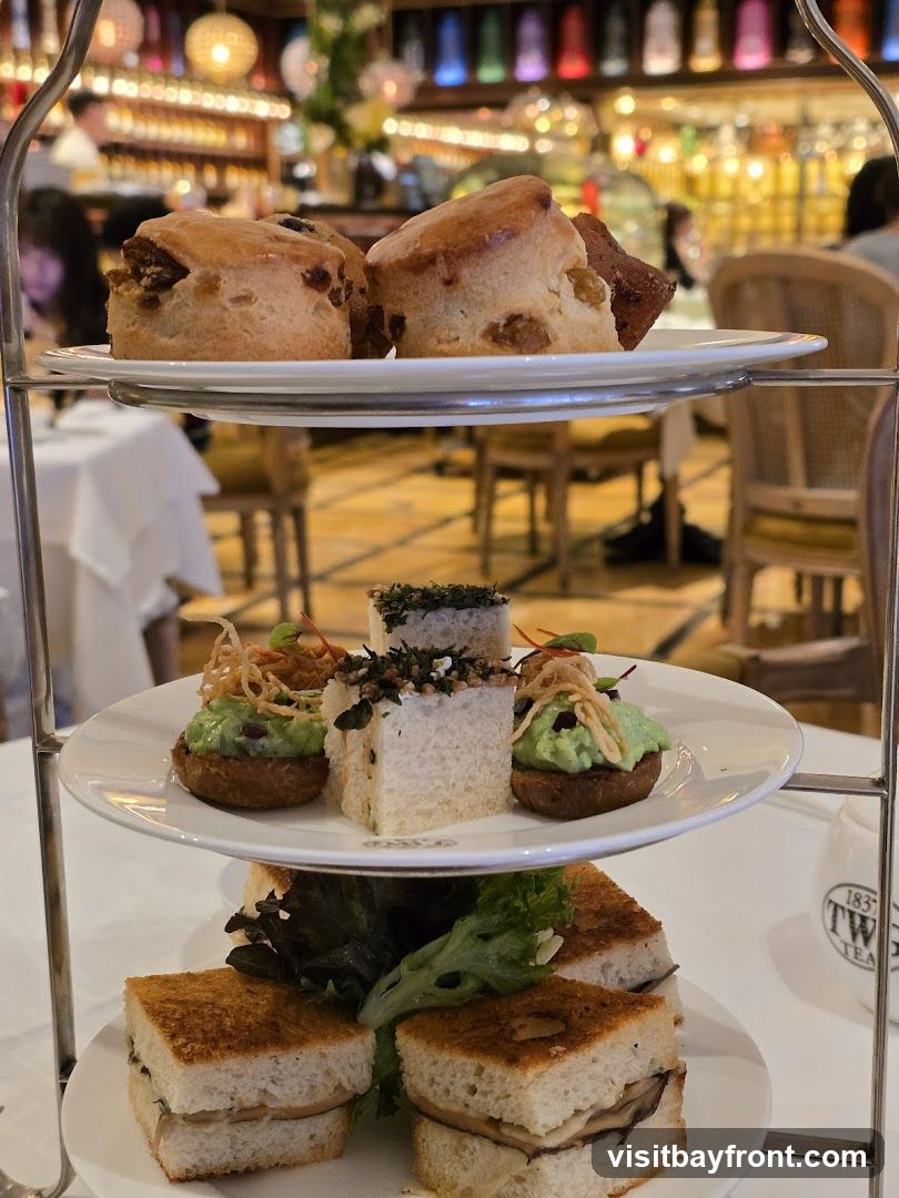 A three-tiered stand holds an assortment of pastries and sandwiches, likely for afternoon tea. The top tier features scones, the middle tier has small savory bites and sandwiches, and the bottom tier displays finger sandwiches. In the background, a restaurant interior with warm lighting and shelves stocked with bottles is visible. A white teapot with the logo "1837 TW... TEA" is partially visible on the right.