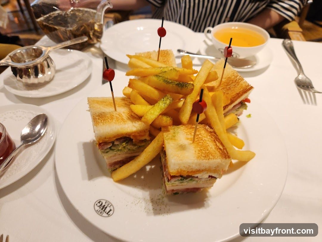 A plate of club sandwiches and french fries is served on a white tablecloth. A teapot filled with tea, a tea strainer, a cup of orange liquid, and a spoon are also visible on the table. The plate has a logo that reads "TWG 1837"