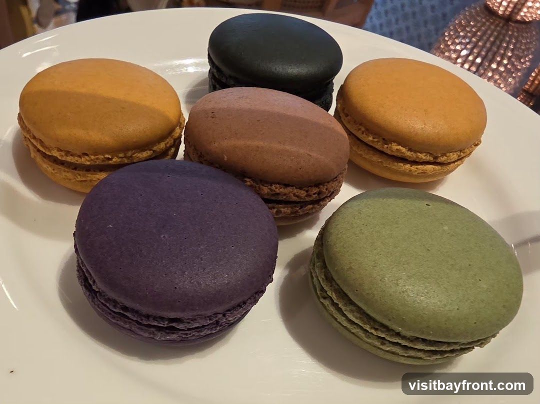 A white plate holds six colorful macarons: two orange, one purple, one brown, one black, and one green.
