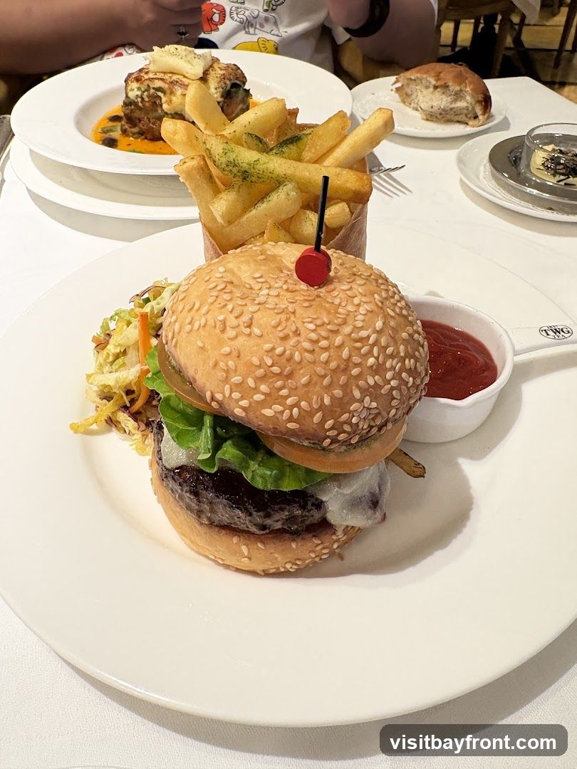 A close-up shot of a gourmet burger served with a side of french fries and coleslaw. The burger is on a sesame seed bun with lettuce, tomato, and cheese. The fries are seasoned with herbs and served in a paper cone. A small dish of ketchup is also visible. In the background, another dish with what appears to be a layered pastry or gratin is seen, along with a bread roll and a small dessert. The setting appears to be a restaurant with white tablecloths. A logo for TWG is visible on a small white dish.
