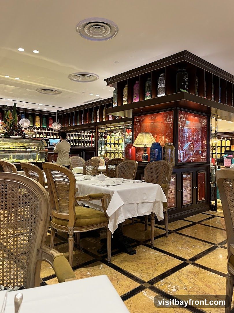 An interior view of a tea room with tables set for service. Wicker-backed chairs with green upholstery surround tables draped in white tablecloths. Shelves filled with decorative tea tins line the walls, and a display case with pastries is visible in the background. A staff member stands behind a counter. The floor is a marble pattern of yellow and black tiles.