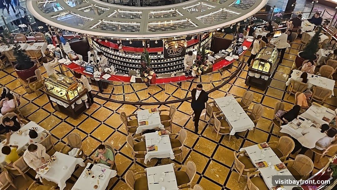 An overhead view of a bustling tea room, featuring a circular bar area with staff serving customers. Tables are set with white tablecloths and chairs, and patrons are seated, enjoying their time. The decor is opulent, with a patterned floor and decorative elements around the bar. A display case with pastries is visible. The TWG Tea logo is prominently displayed on the bar.