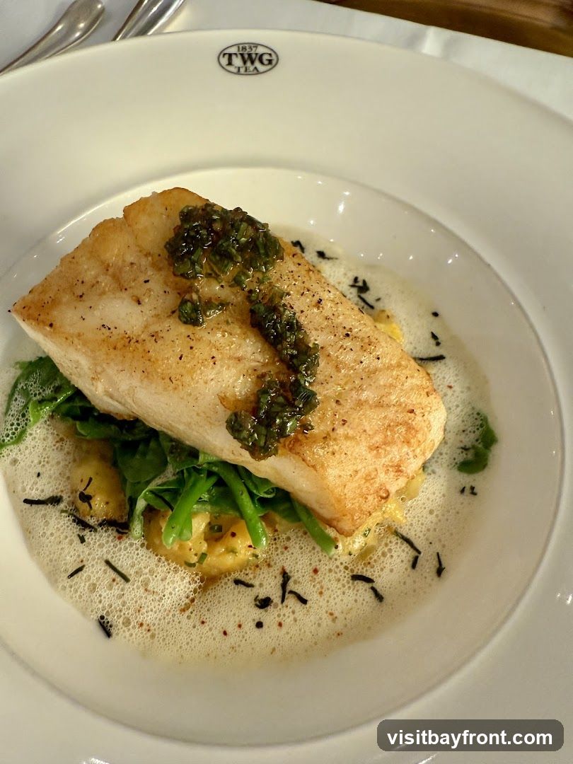 A white plate with a piece of pan-seared fish on top of a bed of greens and a creamy sauce. The fish is topped with a dark green relish. The dish is surrounded by a foamy sauce and garnished with small dark specks. The TWG Tea logo is visible on the rim of the plate.