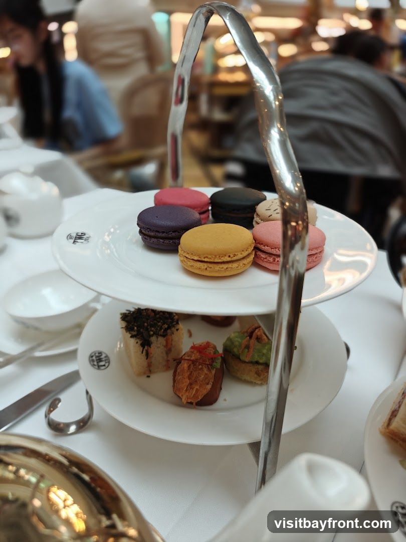 A tiered stand holds an assortment of macarons and small savory bites, likely part of an afternoon tea service. The macarons are in various colors including purple, pink, black, and yellow. The savory items include small sandwiches or canapés topped with ingredients like seaweed, avocado, and shredded meat. The stand has a distinctive metallic handle. In the background, blurred figures and a bright, possibly retail, environment are visible.