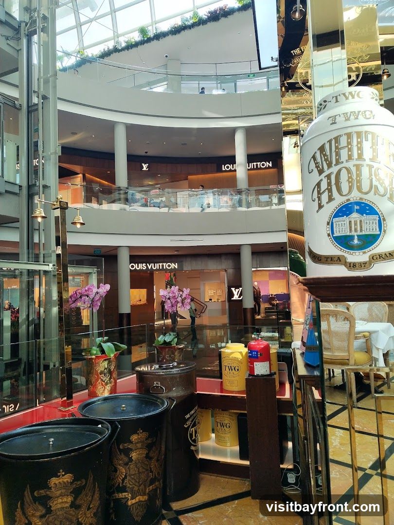 The image shows the interior of a modern shopping mall with multiple levels. A Louis Vuitton store is visible on one of the upper levels, with its signage clearly displayed. In the foreground, there are decorative items, including large ornate containers and canisters labeled TWG Tea. A large tin with "White House TWG Tea 1837" is prominent. The overall impression is of a luxurious retail environment.