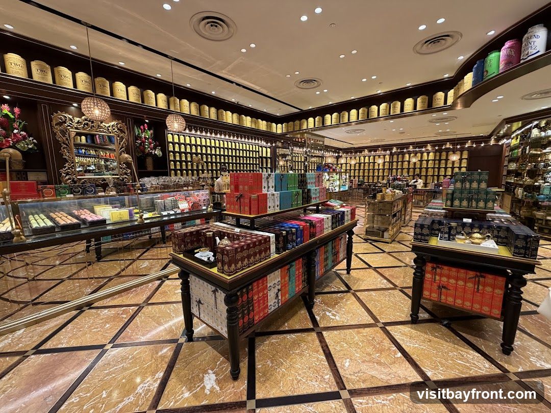 The interior of a well-lit tea shop, featuring numerous shelves stocked with tea tins and boxes. Display tables showcase various tea products, including colorful boxes and decorative teapots. A glass display case in the foreground holds macarons and other confections. The floor is made of polished marble tiles.