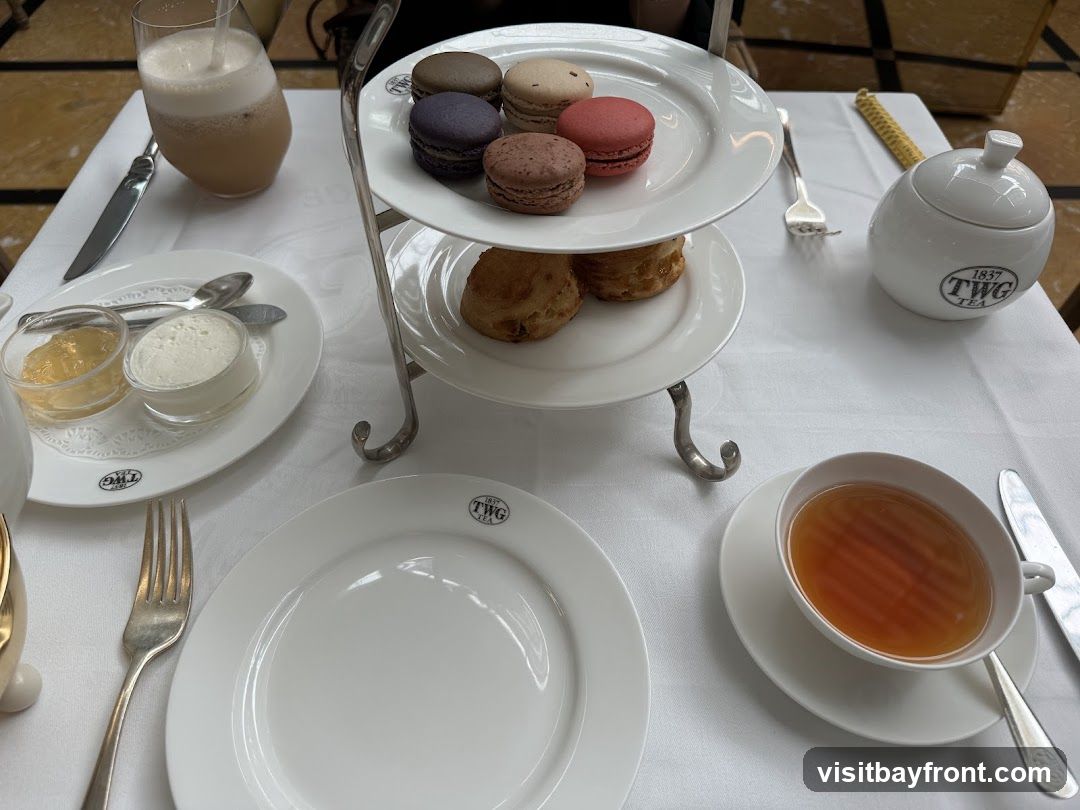 A table set for afternoon tea with a tiered stand holding macarons and scones. A cup of tea, a glass of iced beverage, jam, cream, and plates are also visible. The TWG Tea logo is present on the tableware.
