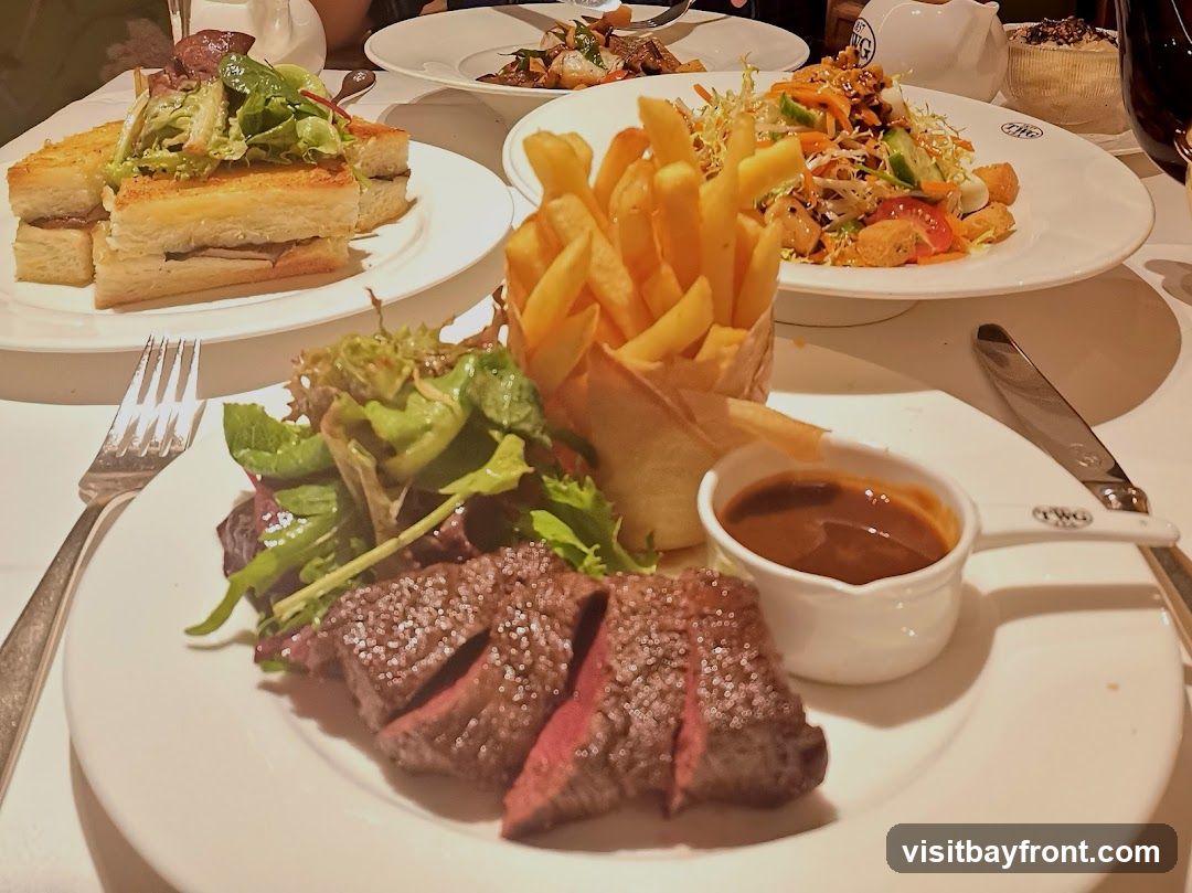 A close-up view of a steak dinner served on a white plate. The steak is sliced and appears medium-rare, accompanied by a side salad and a small dish of gravy. In the background, there are other plates of food, including a sandwich and a salad with fries. The table setting suggests a restaurant environment.