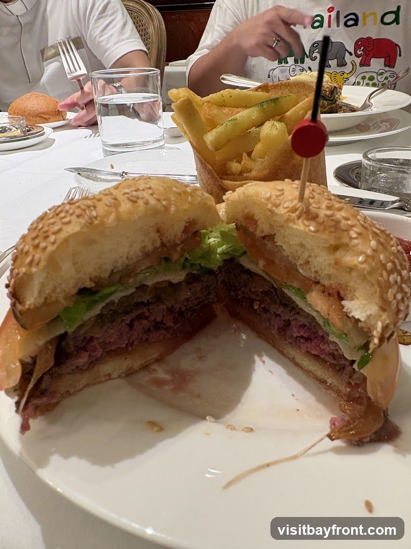 A close-up shot of a halved burger with sesame seed bun, lettuce, tomato, and a thick beef patty, served with a side of french fries in a paper cone. The meal is presented on a white plate, with a glass of water and a bread roll visible in the background.