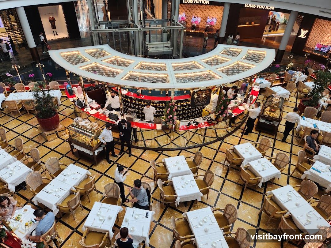 An overhead view of a cafe situated in a shopping mall. The cafe features a curved counter with staff serving customers. Tables are set with white tablecloths and chairs are arranged around them, with some patrons seated and dining. In the background, high-end retail stores like Louis Vuitton are visible.