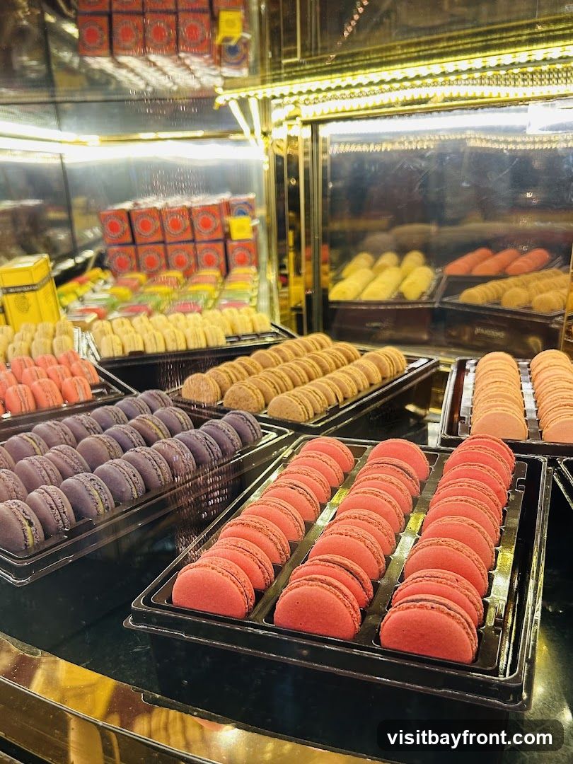 A display case filled with rows of colorful macarons. In the foreground, trays of pink and purple macarons are visible. Behind them, rows of beige and yellow macarons are arranged. In the background, boxes and other pastries are displayed under bright lighting.