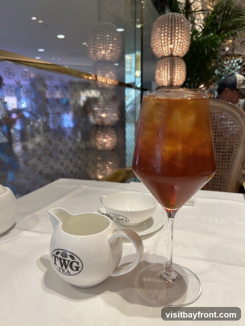 A tall glass of iced tea sits on a white tablecloth next to a small creamer pitcher. Both the glass and the pitcher have the TWG Tea logo. In the background, a white teacup and saucer are visible, also with the TWG Tea logo. Decorative lamps and greenery are also present in the background, suggesting an indoor dining setting.