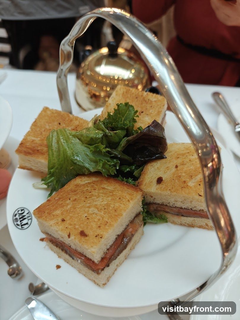 A tiered stand holds several small, square sandwiches with toasted bread and a filling of meat and some sort of relish or spread. A small garnish of green lettuce is placed between the sandwiches. In the background, a golden teapot and other tableware are visible. The TWG Tea logo is visible on the plate.