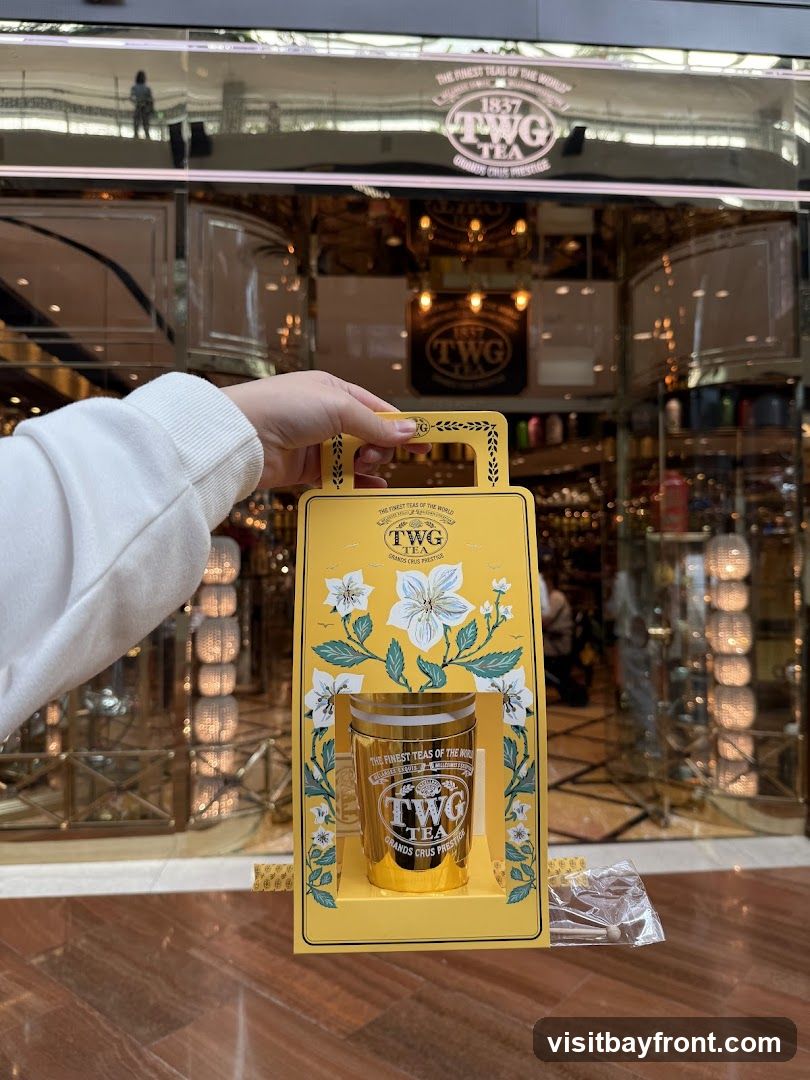 A hand holds a yellow gift box containing a gold tin of TWG tea. The background shows the exterior of a TWG Tea shop with its signage visible.