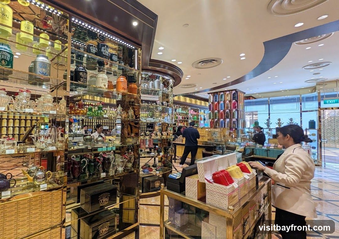 A well-lit interior of a luxury tea shop, showcasing an extensive collection of teas in decorative tins and jars displayed on mirrored shelves. Various teapots, cups, and other tea accessories are also visible. Customers and staff are browsing and interacting within the store.