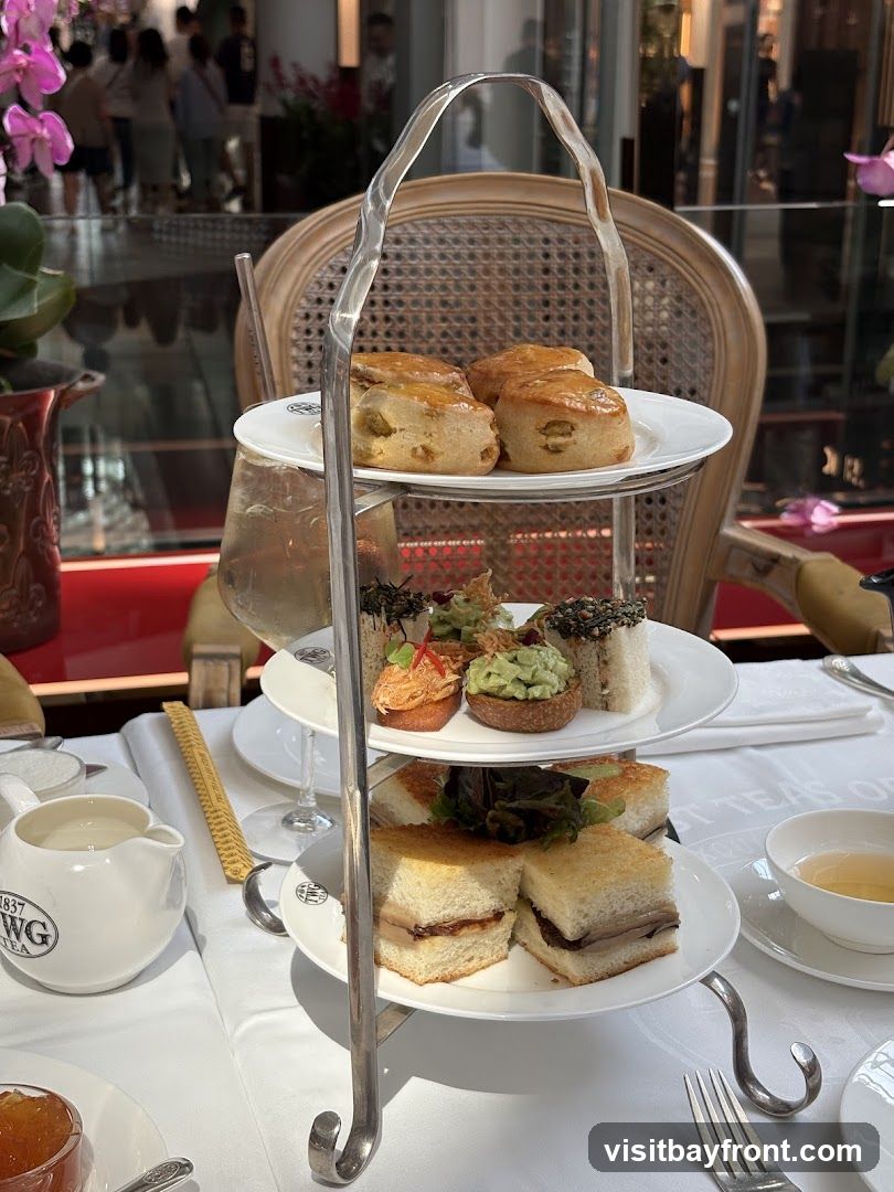 A three-tiered stand holds an assortment of pastries, sandwiches, and scones, set on a white tablecloth. A pitcher with the logo "1837 TWG TEA" is visible on the left. In the background, blurred figures are seen walking past a storefront.