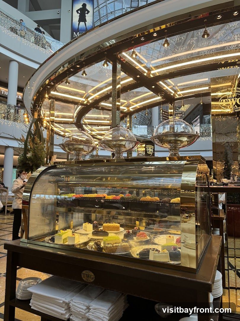 A display case filled with various cakes and pastries, including macarons, layered cakes, and tarts. The display is illuminated by overhead lights and reflects the surrounding mall environment. A person in a white uniform and mask is visible in the background.