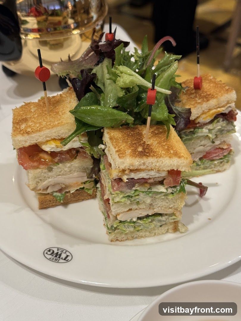 Three club sandwiches are presented on a white plate with a side of mixed greens salad. The sandwiches are cut into quarters and held together with toothpicks. The plate has a logo that reads "TWG TEA 1837" in a circular emblem. A silver teapot is visible in the blurred background.