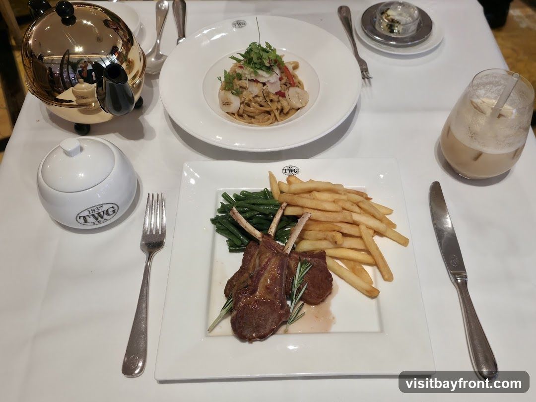 A table setting with a meal of lamb chops, french fries, and green beans on a white plate. Another plate holds a pasta dish with seafood. A teapot, sugar bowl, and a drink are also on the table. The TWG Tea logo is visible on the sugar bowl and the plate.