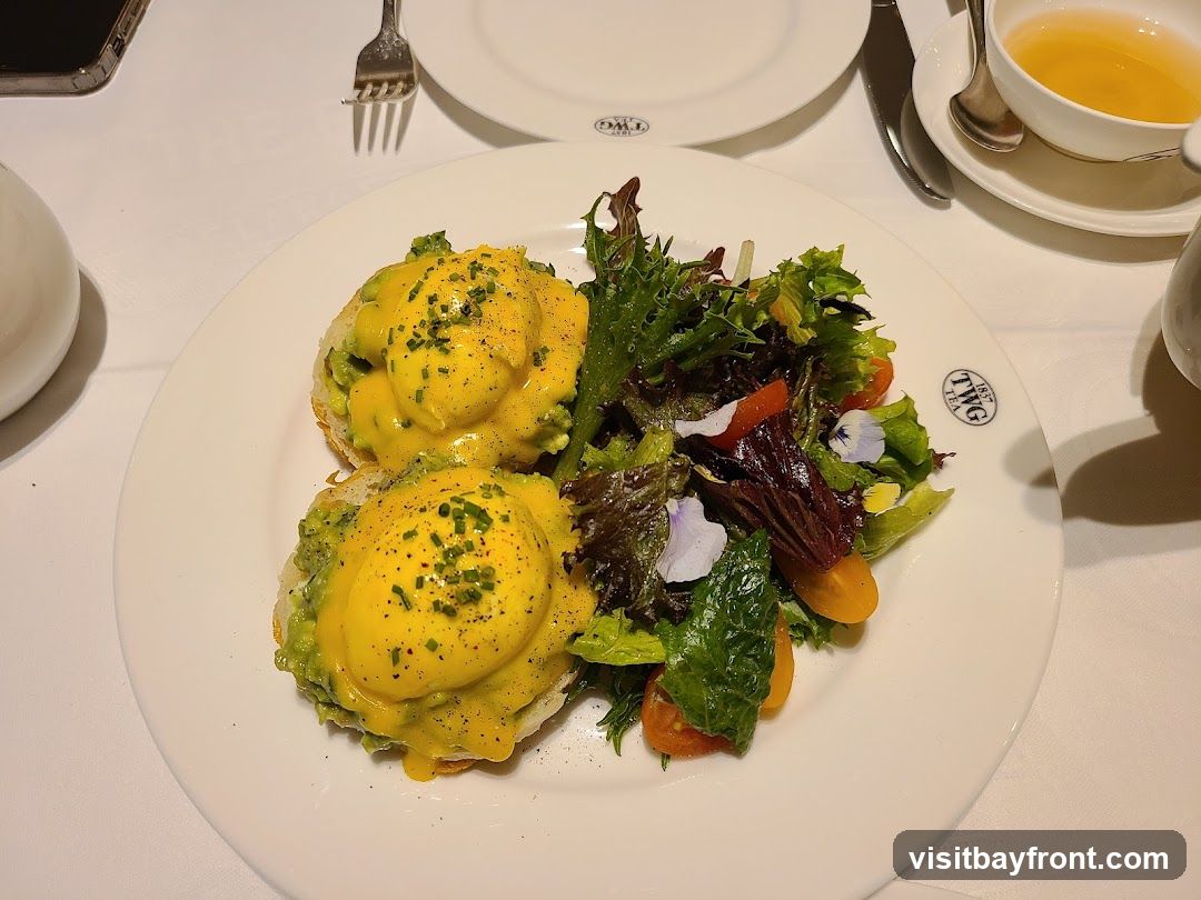 A plate of Eggs Benedict served with a side salad. The dish consists of two halves of an English muffin, each topped with avocado, poached eggs, and hollandaise sauce, garnished with chives and black pepper. The salad is a mix of greens, cherry tomatoes, and edible flowers. A cup of tea and a phone are also visible on the table. The plate has a TWG 1837 Tea logo.