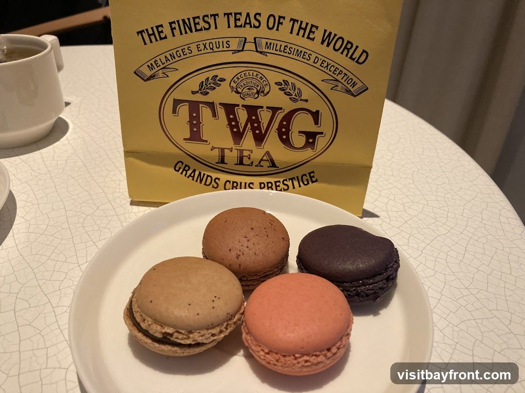 A plate of four macarons sits on a white table next to a yellow TWG Tea bag and a cup of tea. The macarons are in various colors: brown, light brown, pink, and dark purple. The TWG Tea bag has the brand's logo and text indicating "The Finest Teas of the World."
