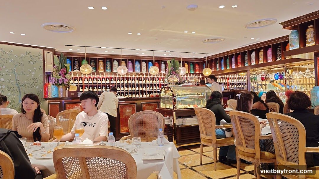 A well-lit restaurant interior with several patrons seated at tables. The walls are lined with shelves displaying numerous colorful bottles and what appear to be tea tins. A display case with pastries is visible, along with a bar area stocked with bottles. The decor includes floral wallpaper and ornate lighting fixtures.