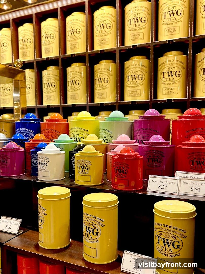 A display of colorful TWG tea tins arranged on shelves. The tins are of various sizes and colors, including yellow, red, pink, purple, green, blue, and white. Some tins have labels indicating the tea type and weight, with prices visible for some items.