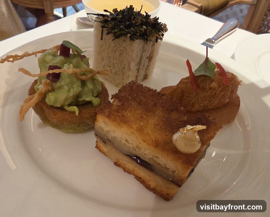 A white plate holds three small savory appetizers. On the left is a tartlet filled with mashed avocado, topped with crispy strands and diced red garnish. In the center, a small sandwich is topped with dark green herbs. On the right, a crispy, golden-brown item is garnished with red slivers and a small green leaf. A slice of layered food, possibly a sandwich or pastry, with a dark filling and a golden leaf garnish, is in the foreground. A bowl of yellow soup is visible in the background.