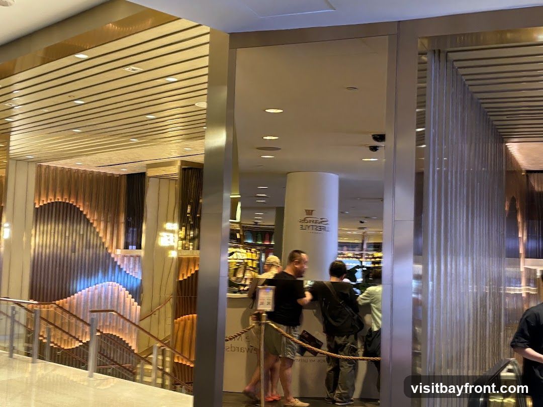 The image shows the interior of a store with a modern design. There are several people standing around a counter, and a staircase with decorative lighting is visible on the left. The ceiling has a slatted design, and there are mirrors and metallic accents throughout the space.