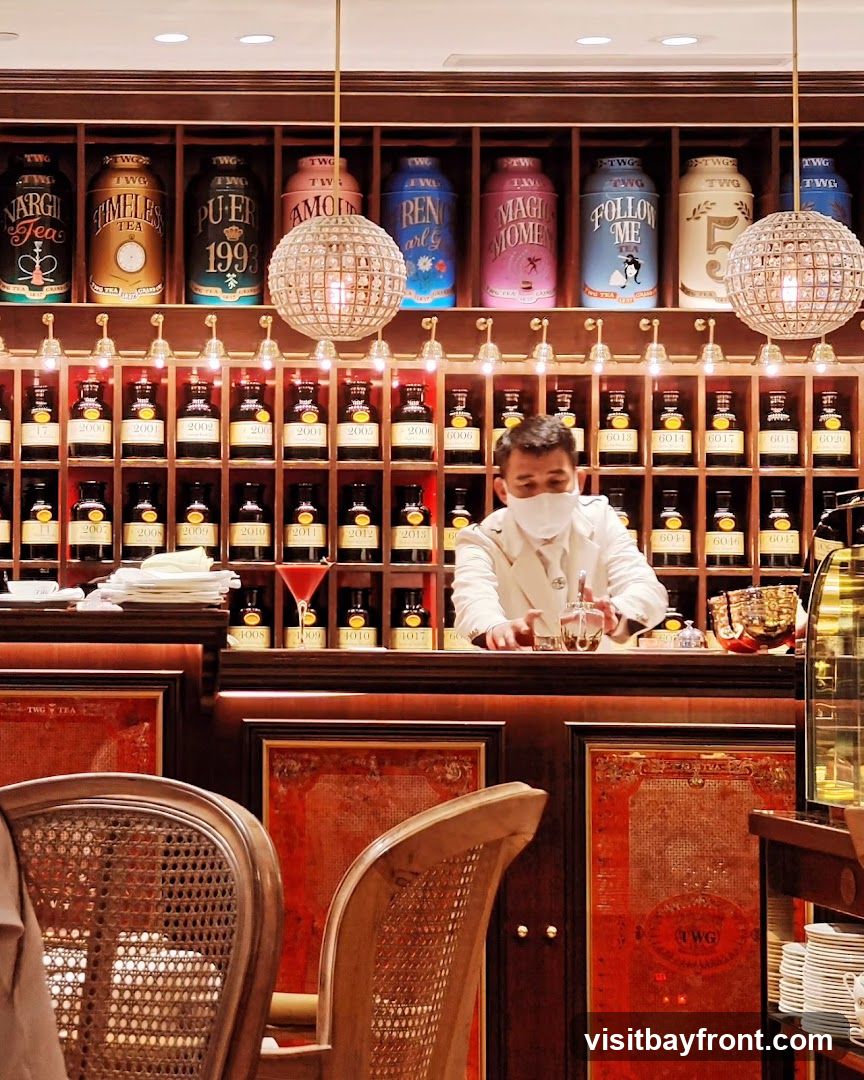 A man wearing a mask and a white coat stands behind a counter in a tea shop. He is preparing a drink. The shelves behind him are filled with numerous tins of tea and bottles. The decor is ornate with red accents and chandeliers.