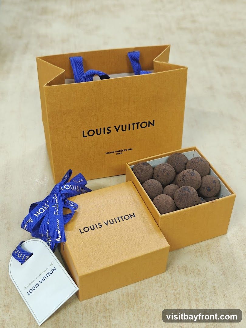 A Louis Vuitton shopping bag, a small Louis Vuitton box, and a box of chocolates are arranged on a light-colored surface. The shopping bag and box are branded with the Louis Vuitton logo. A blue ribbon with the Louis Vuitton logo is tied around the small box. A tag with "Maxime Frederic of LOUIS VUITTON" is attached to the ribbon. The box of chocolates contains several round, dark brown chocolates dusted with cocoa powder.