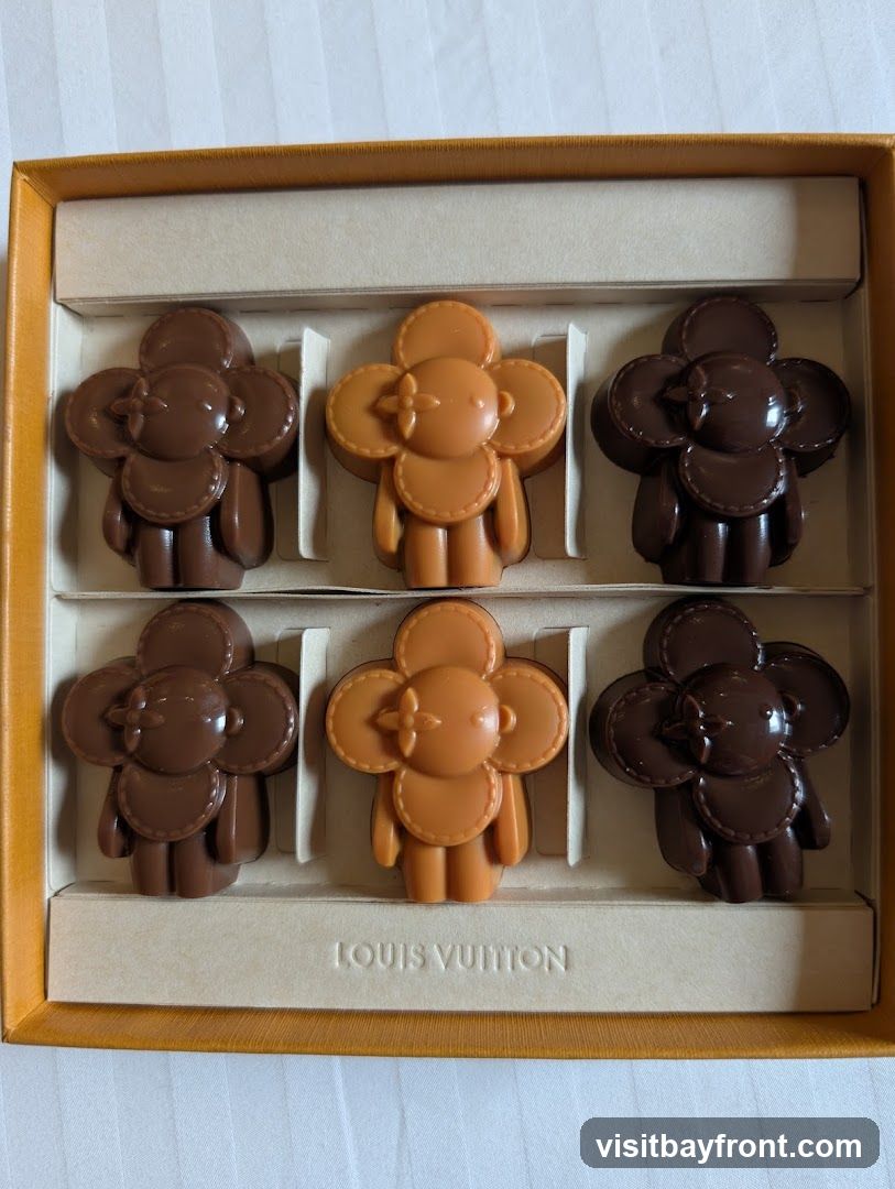 A box of six chocolates shaped like flowers with faces, arranged in two rows of three. The chocolates are in two colors: dark brown and a lighter caramel brown. The box is orange with a cream-colored insert that has the embossed text "LOUIS VUITTON" at the bottom.