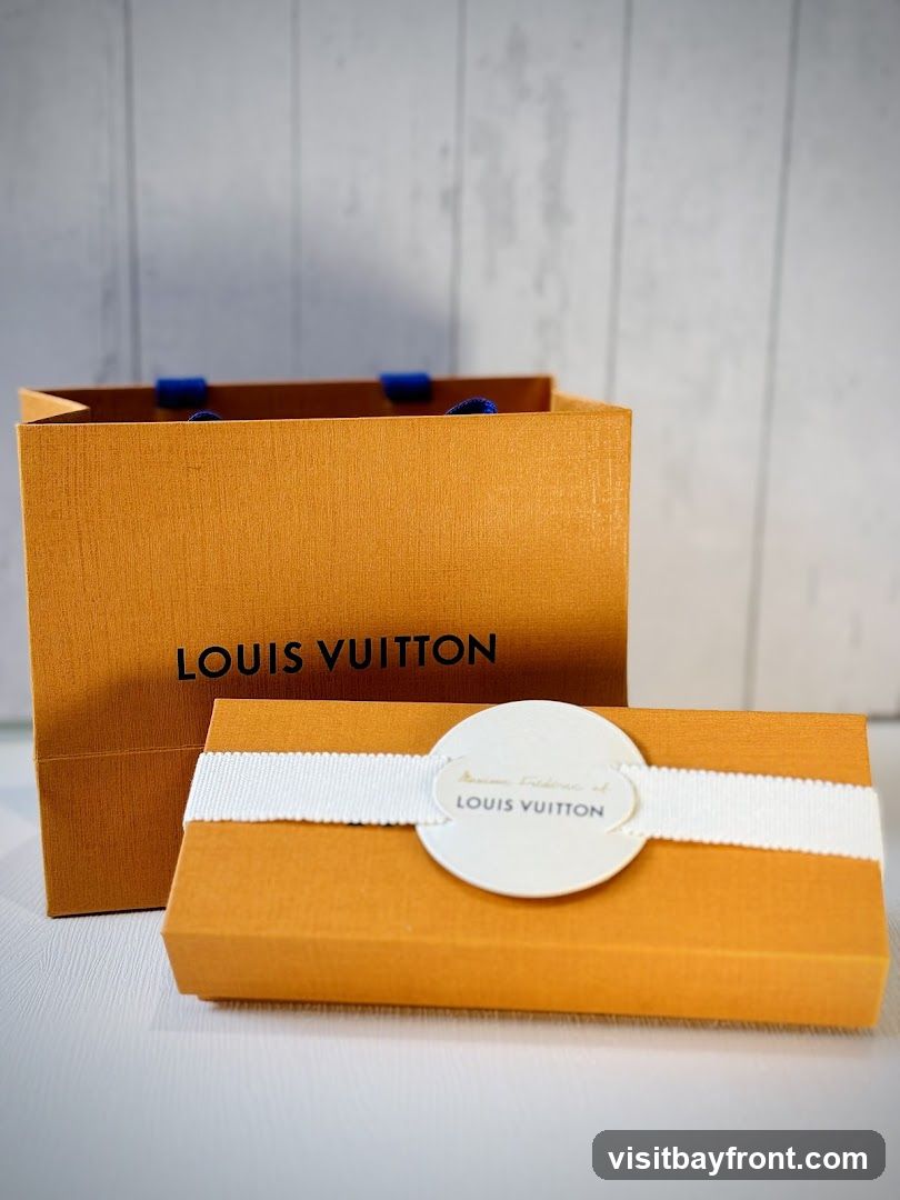 A close-up shot of a Louis Vuitton shopping bag and a gift box. The orange bag has the Louis Vuitton logo printed in black. The orange gift box is tied with a white ribbon and has a circular tag with text that includes "LOUIS VUITTON" and "Maison Fréderic et". The background is a light-colored, textured wall.