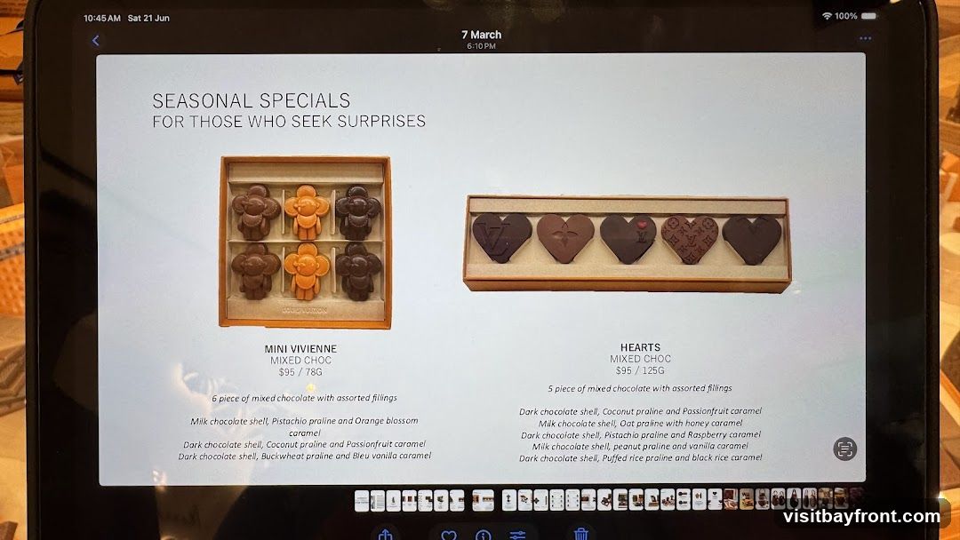 A tablet screen displaying seasonal chocolate specials. Two boxes of chocolates are shown: 'MINI VIVIENNE MIXED CHOC' (6 pieces for $95/78G) and 'HEARTS MIXED CHOC' (5 pieces for $95/125G). Descriptions of the chocolate shells and fillings are provided for each.