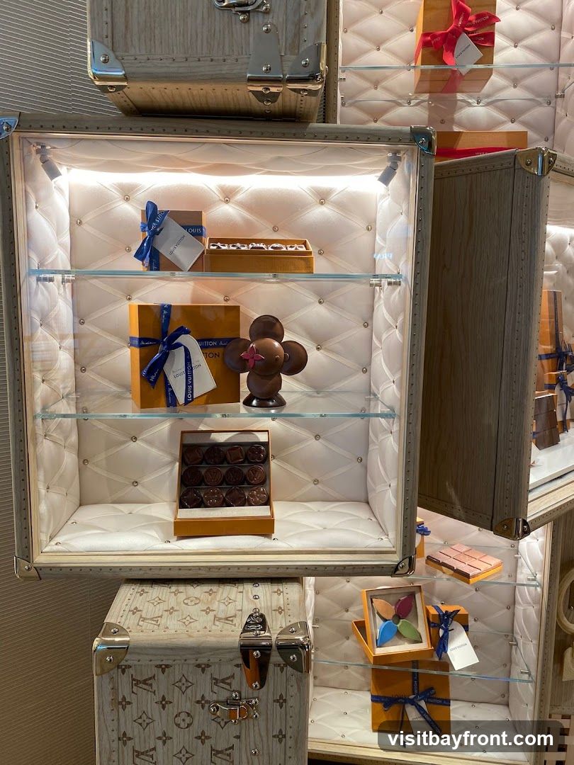 A display case filled with Louis Vuitton branded chocolates and gift boxes. The chocolates are arranged in trays and some are shaped like the LV flower. The display is set within a trunk-like structure with quilted white lining and illuminated shelves.