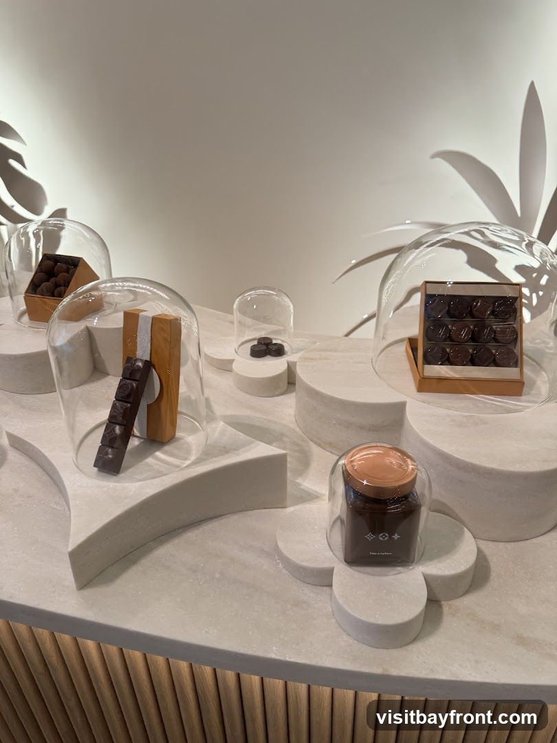 A display of various chocolates under glass domes on a textured surface. Some chocolates are in boxes, while others are loose. A jar with a lid and a logo is also visible. The background is a plain wall with abstract leaf-like shapes.