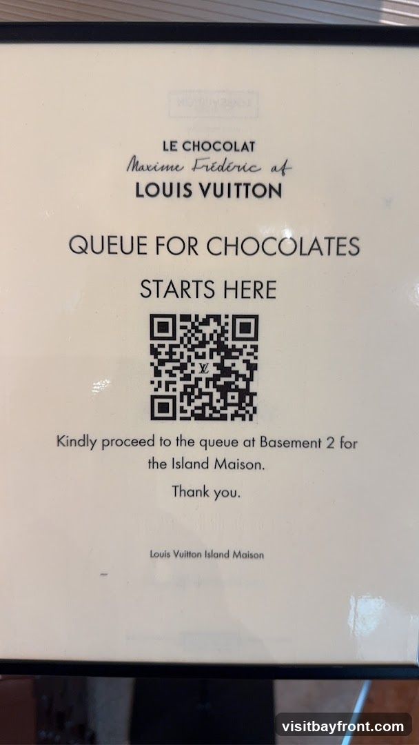 A sign indicating where the queue for chocolates starts. It reads 'LE CHOCOLAT Maxime Frédéric at LOUIS VUITTON QUEUE FOR CHOCOLATES STARTS HERE' with a QR code. Below the QR code, it instructs to 'Kindly proceed to the queue at Basement 2 for the Island Maison. Thank you.' The sign also displays 'Louis Vuitton Island Maison' at the bottom.