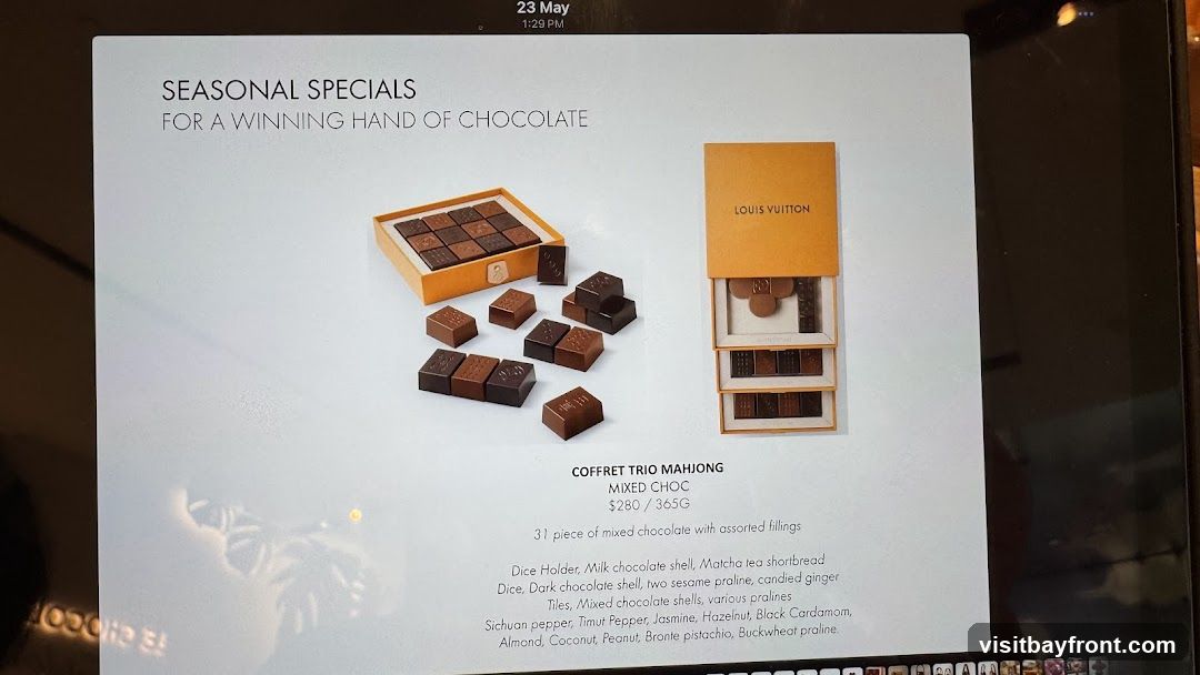 A display of Louis Vuitton chocolates, featuring a box labeled "COFFRET TRIO MAHJONG MIXED CHOC" priced at $280 for 365g. The description lists 31 pieces of mixed chocolate with assorted fillings, including various praline flavors and ingredients like matcha tea shortbread, candied ginger, Sichuan pepper, and hazelnut.