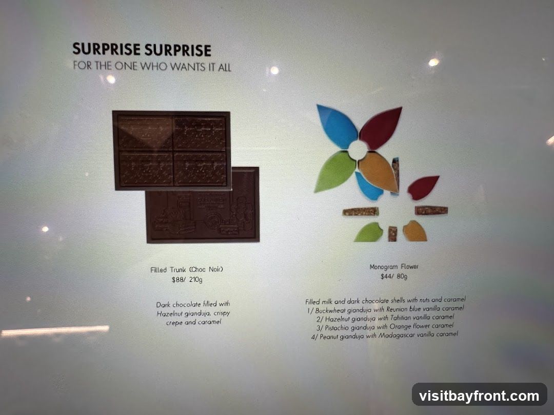 Two chocolate products are displayed: 'Filled Trunk (Choc Noir)' and 'Monogram Flower'. The Filled Trunk is described as dark chocolate filled with hazelnut gianduja, crispy crepe, and caramel, priced at $88 for 210g. The Monogram Flower consists of filled milk and dark chocolate shells with nuts and caramel, with four flavor options: Buckwheat gianduja with Reunion blue vanilla caramel, Hazelnut gianduja with Tahitian vanilla caramel, Pistachio gianduja with Orange flower caramel, and Peanut gianduja with Madagascar vanilla caramel, priced at $44 for 80g.