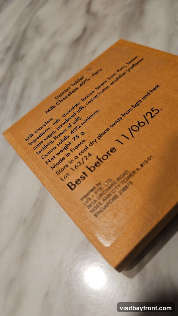 The image shows the back of an orange-colored packaging for a milk chocolate bar. It lists the product as 'Damier Tablet Milk Chocolate 40% - Peru'. The ingredients include milk chocolate, cane sugar, powdered milk, cocoa butter, sunflower lecithin, and flower of salt. It states the cocoa solids are a minimum of 40%, the net weight is 75g, and it was made in France. The packaging also includes storage instructions and a 'Best before' date of 11/06/25. Import details for LVS PTE LTD in Singapore are also visible.