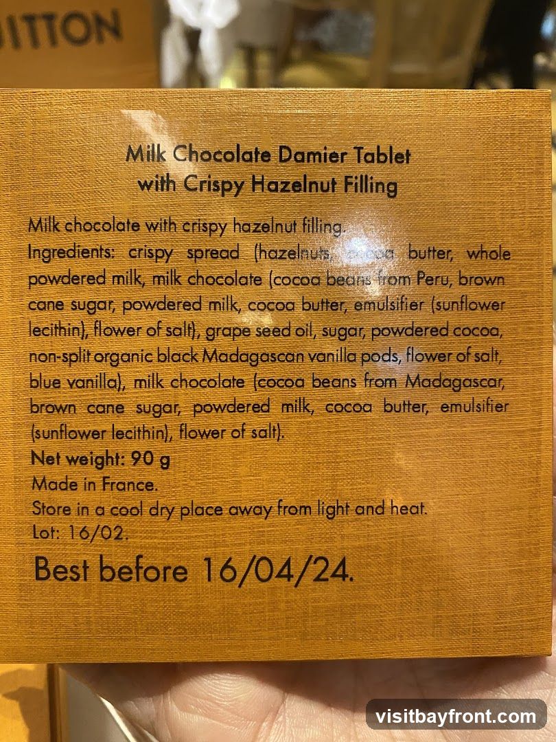 The image shows the packaging of a milk chocolate tablet with crispy hazelnut filling. The text details the ingredients, net weight, origin (Made in France), storage instructions, lot number, and best before date. The packaging has a textured, wood-like finish.