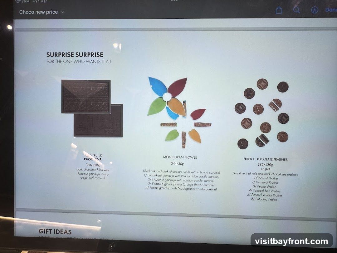 A digital display showing gift ideas for chocolates. It features three items: Filled Trunk Choco Noir, Monogram Flower, and Filled Chocolate Pralines. Each item has a description, price, and weight.